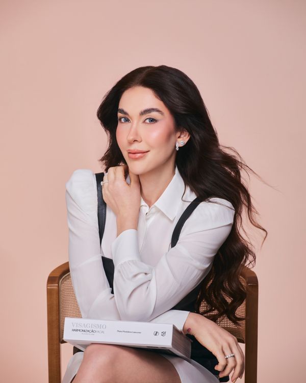 Dra. Katarina Bessa sentada, de camisa branca, em fundo rosé